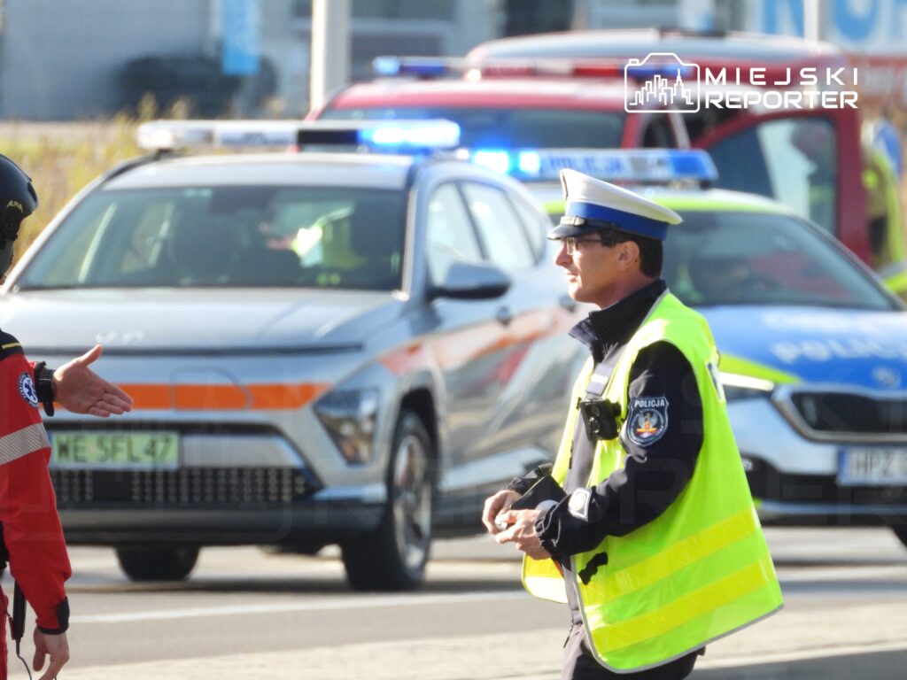 Policjant w żółtej kamizelce kieruje ruchem drogowym obok radiowozów na ulicy w mieście.