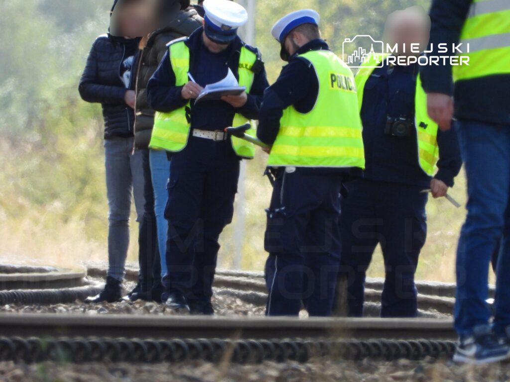 Policjanci w odblaskowych kamizelkach prowadzą rozmowę i spisują notatki na torach kolejowych.