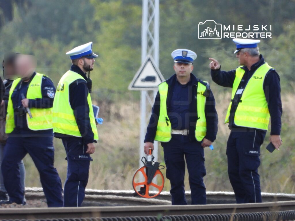 Grupa policjantów w kamizelkach odblaskowych dyskutuje na torach kolejowych, jeden z nich trzyma urządzenie pomiarowe.