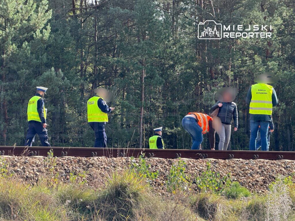 Policjanci w odblaskowych kamizelkach prowadzą czynności na torach kolejowych, w otoczeniu drzew.