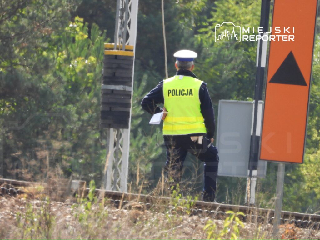 Policjant w kamizelce odblaskowej stojący obok torów kolejowych, trzymający dokumenty w ręku.