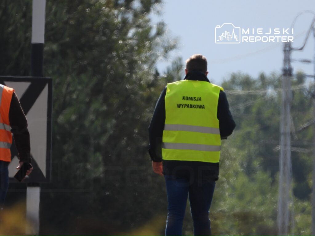 Mężczyzna w kamizelce odblaskowej z napisem "KOMISJA WYPADKOWA" idzie wzdłuż drogi w otoczeniu drzew.