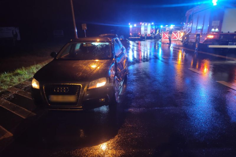 Czarny samochód osobowy stoi na mokrej drodze, obok widać pojazdy straży pożarnej z włączonymi sygnalizacjami.