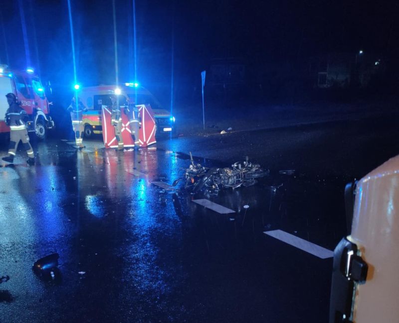 Na drodze w nocy leży uszkodzony motocykl, obok pracują strażacy i karetka z włączonymi sygnalizatorami.