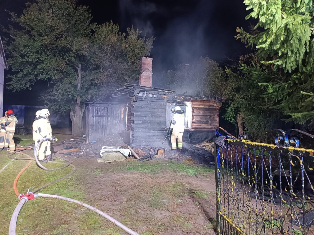 Strażacy gaszą pożar drewnianego budynku w nocy, otoczeni dymem i zniszczeniami na terenie posesji.