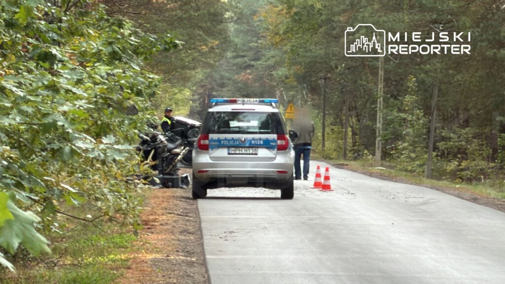 Policyjny radiowóz zatrzymany przy drodze, obok leży motocykl, a funkcjonariusze zabezpieczają miejsce zdarzenia.