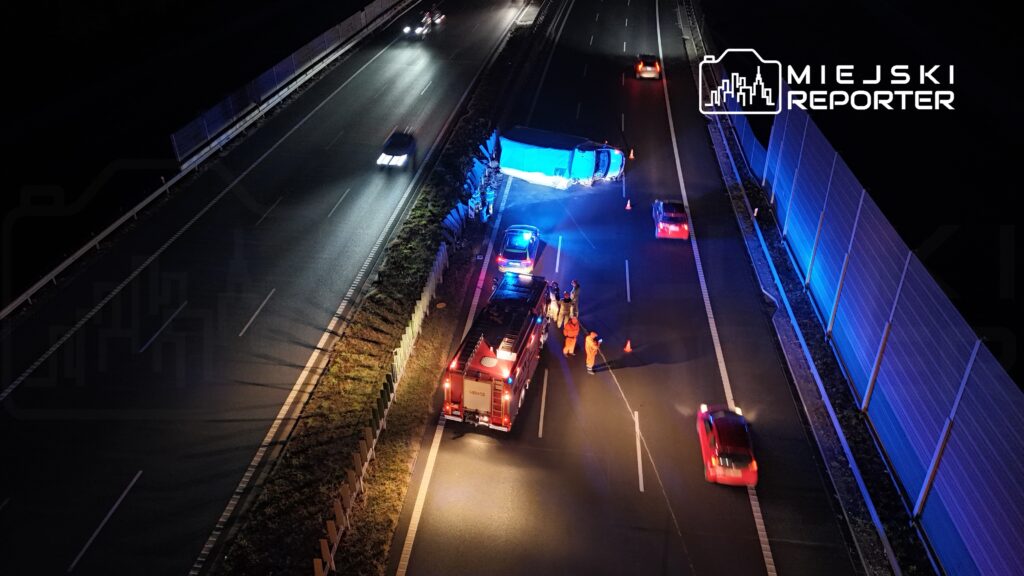 Na drodze leży przewrócony niebieski pojazd, a obok pracują strażacy w pomarańczowych strojach.