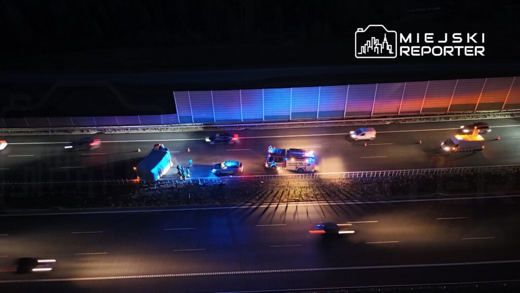 Na drodze leży przewrócony pojazd, a obok pracują służby ratunkowe z włączonymi sygnalizacjami świetlnymi.
