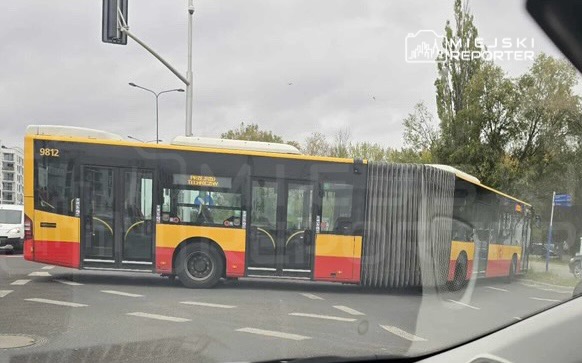 Autobus przegubowy skręca na skrzyżowaniu, otoczony przez inne pojazdy i zieleń w tle.