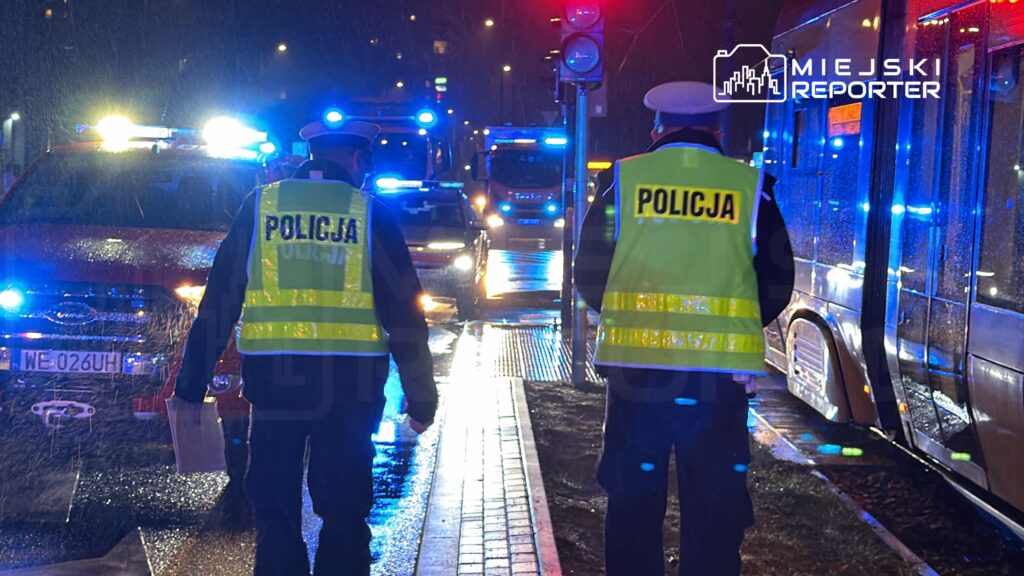 Dwaj policjanci w odblaskowych kamizelkach kierują ruchem na mokrej ulicy, obok zaparkowanego wozu strażackiego.