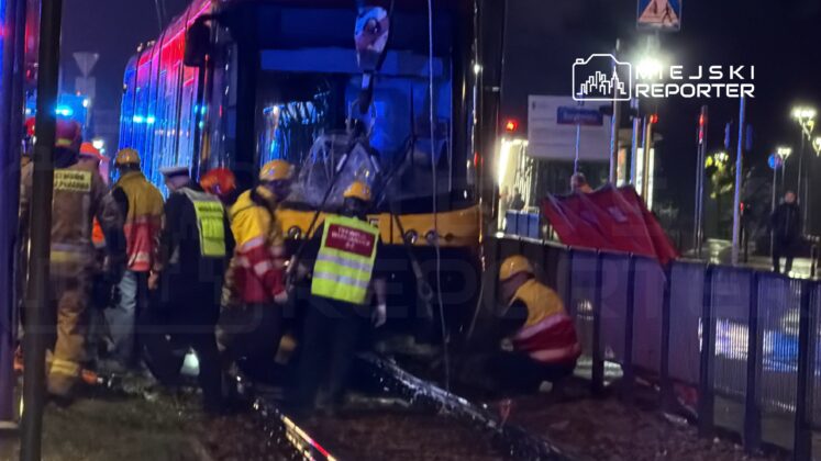 Grupa ratowników w kaskach pracuje przy tramwaju, w tle migają niebieskie światła radiowozów, nocna sceneria.