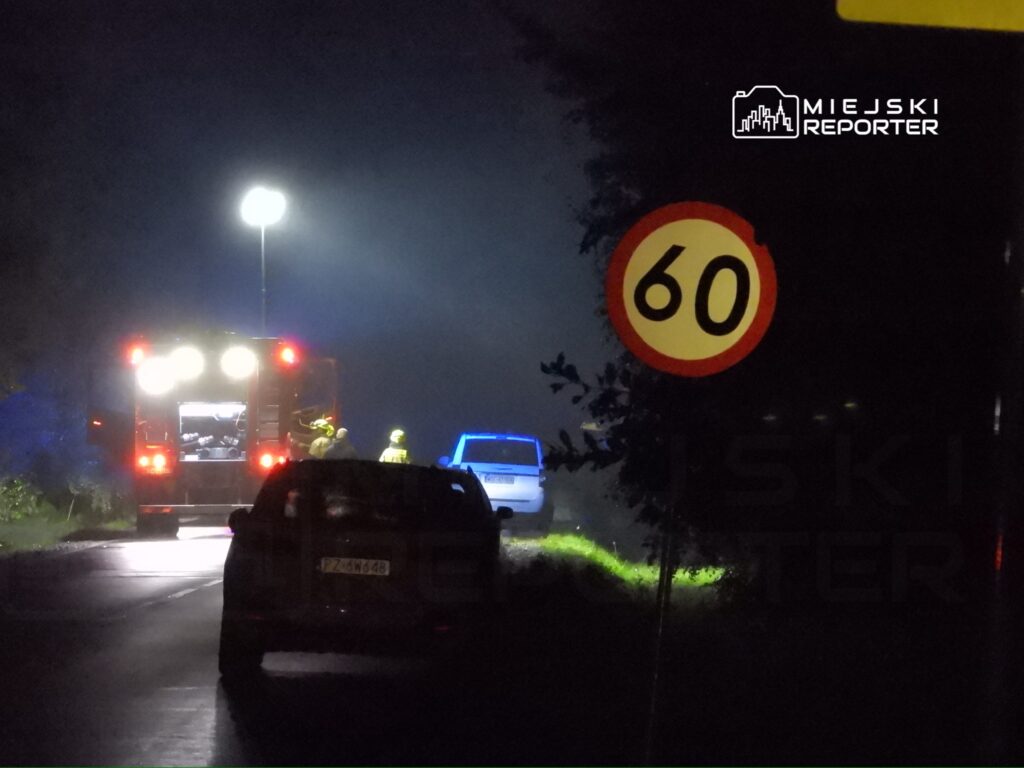 Na drodze w nocy, strażacy przy wozie strażackim prowadzą działania ratunkowe obok zaparkowanego pojazdu.