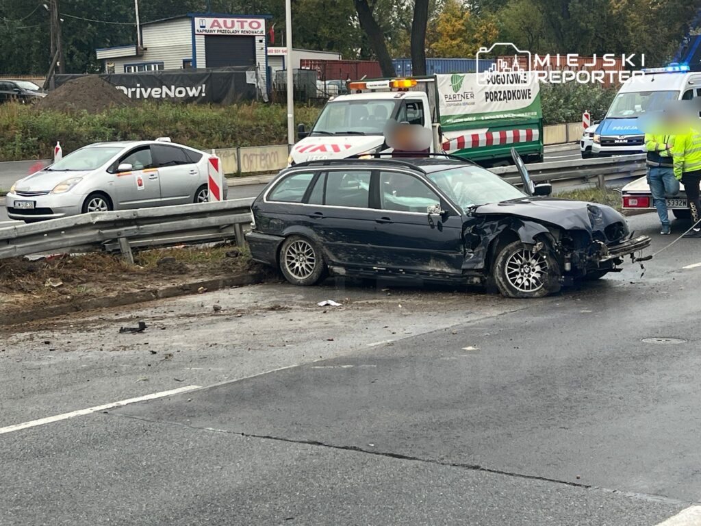 Czarny samochód osobowy z uszkodzeniami stoi przy barierce na drodze, obok interweniujący policjanci i służby.