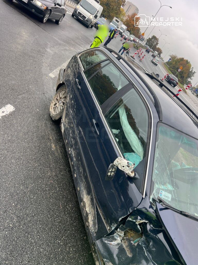 Czarny samochód osobowy z uszkodzeniem bocznej części stoi na drodze, obok pracujący policjanci i inne pojazdy.