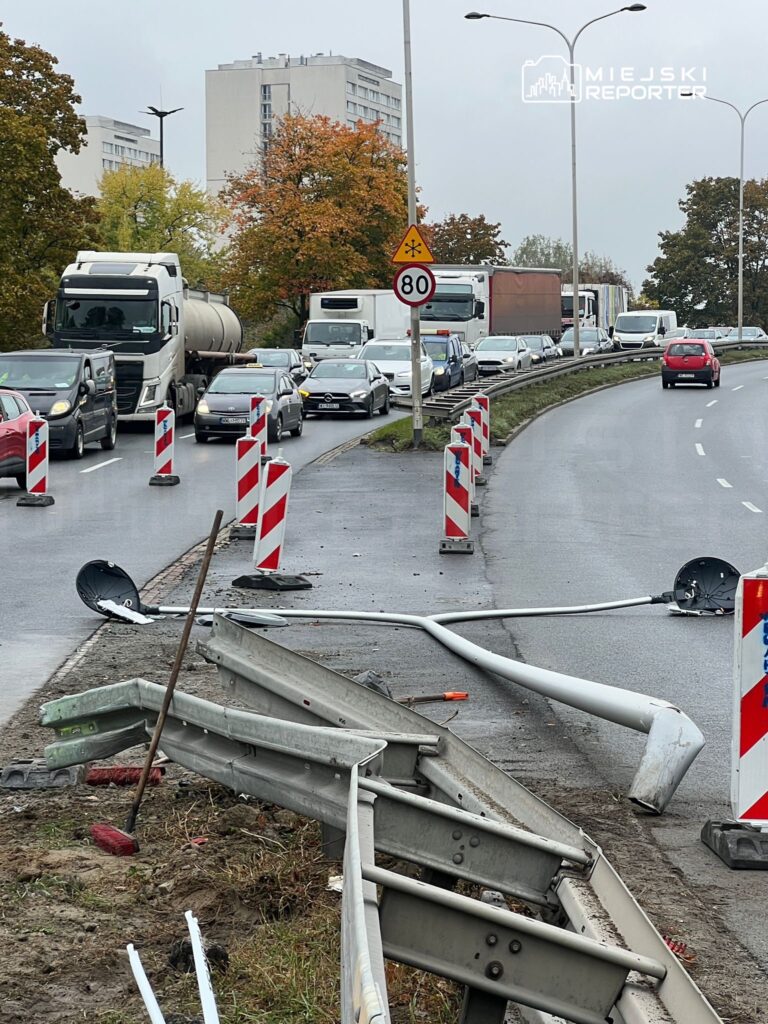 Korki na drodze z uszkodzoną barierką ochronną i powalonym słupem oświetleniowym w tle budynków mieszkalnych.