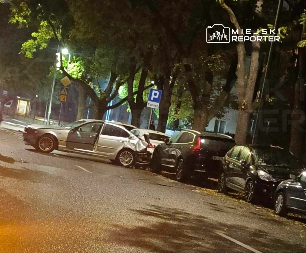 Uszkodzony biały samochód z otwartymi drzwiami stoi na ulicy obok zaparkowanych pojazdów, w tle widoczne drzewa.