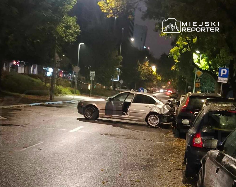 Zniszczony samochód osobowy stoi na ulicy, otoczony innymi zaparkowanymi pojazdami, w nocy.
