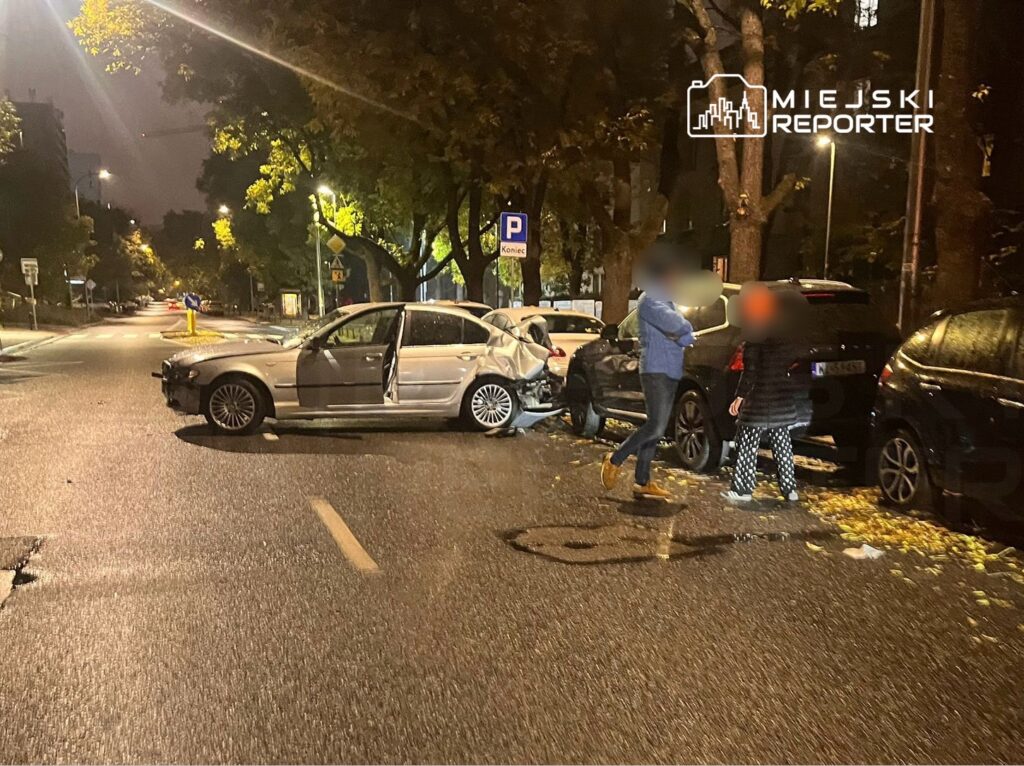 Uszkodzony samochód osobowy z otwartymi drzwiami stoi na ulicy, obok dwóch osób rozmawiających w nocy.