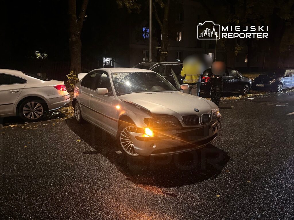 Uszkodzony samochód osobowy marki BMW zaparkowany na deszczowej ulicy, obok innych pojazdów.