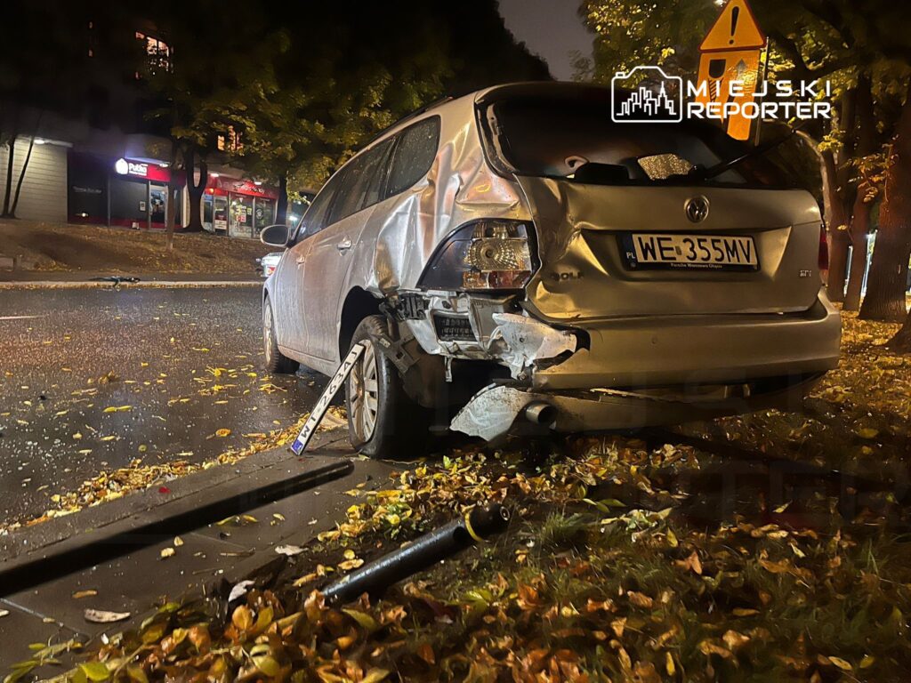 Uszkodzony samochód osobowy zaparkowany na ulicy, otoczony opadłymi liśćmi, w tle widoczne oświetlone sklepy.