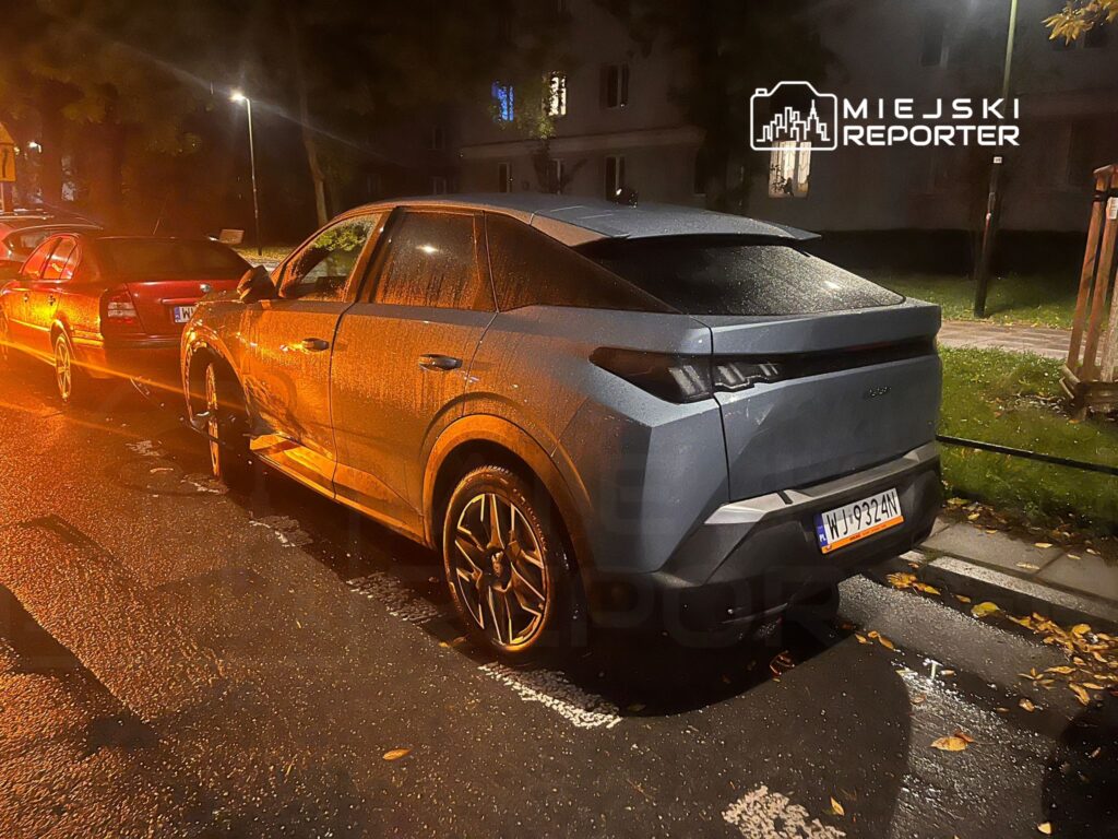 Nowoczesny samochód elektryczny zaparkowany na ulicy w deszczową noc, obok innych pojazdów.