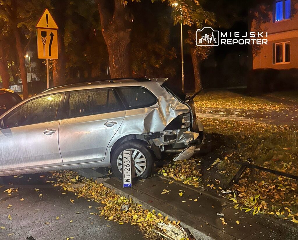 Uszkodzony samochód osobowy zaparkowany na chodniku, obok leżące liście, w tle latarnia i znak drogowy.