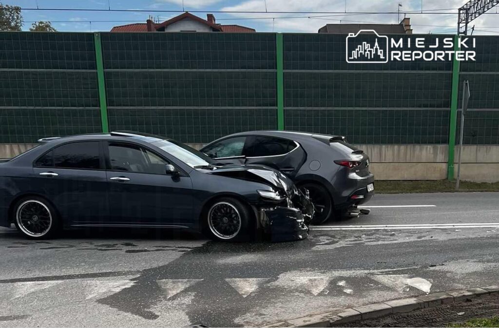Dwa uszkodzone samochody osobowe stoją na jezdni, jeden z nich z wgniecionym przodem. W tle widoczny ogrodzenie i niebieskie niebo.