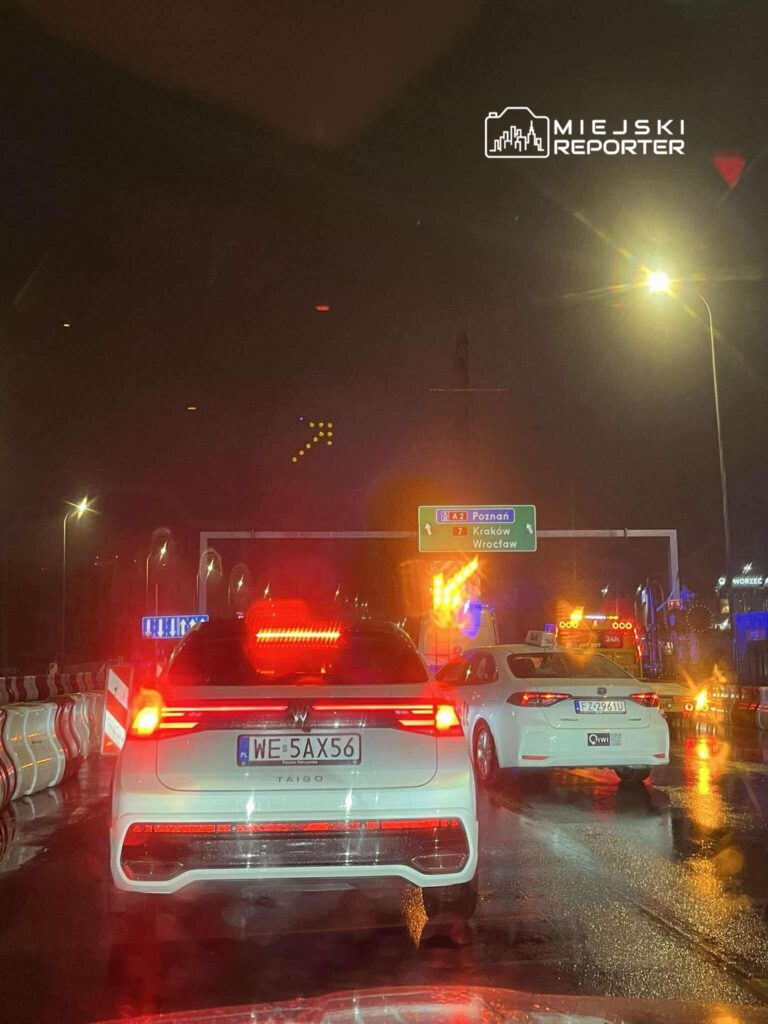 Ruch samochodowy na mokrej drodze w nocy, z widocznymi światłami pojazdów i znakami drogowymi.