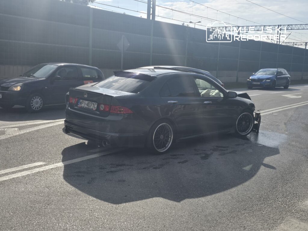 Czarny sedan z uszkodzoną tylną częścią stoi na ulicy, obok przejeżdżają inne samochody.