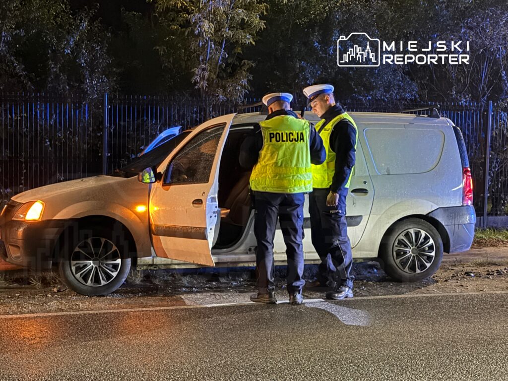 Dwaj policjanci w odblaskowych kamizelkach rozmawiają obok zatrzymanego samochodu na drodze w nocy.