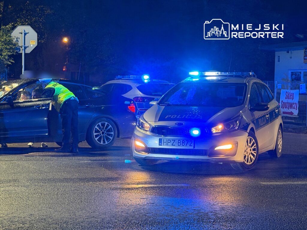 Policjant w kamizelce odblaskowej rozmawia z kierowcą w ciemnej ulicy, obok stoją policyjne radiowozy z włączonymi sygnałami.