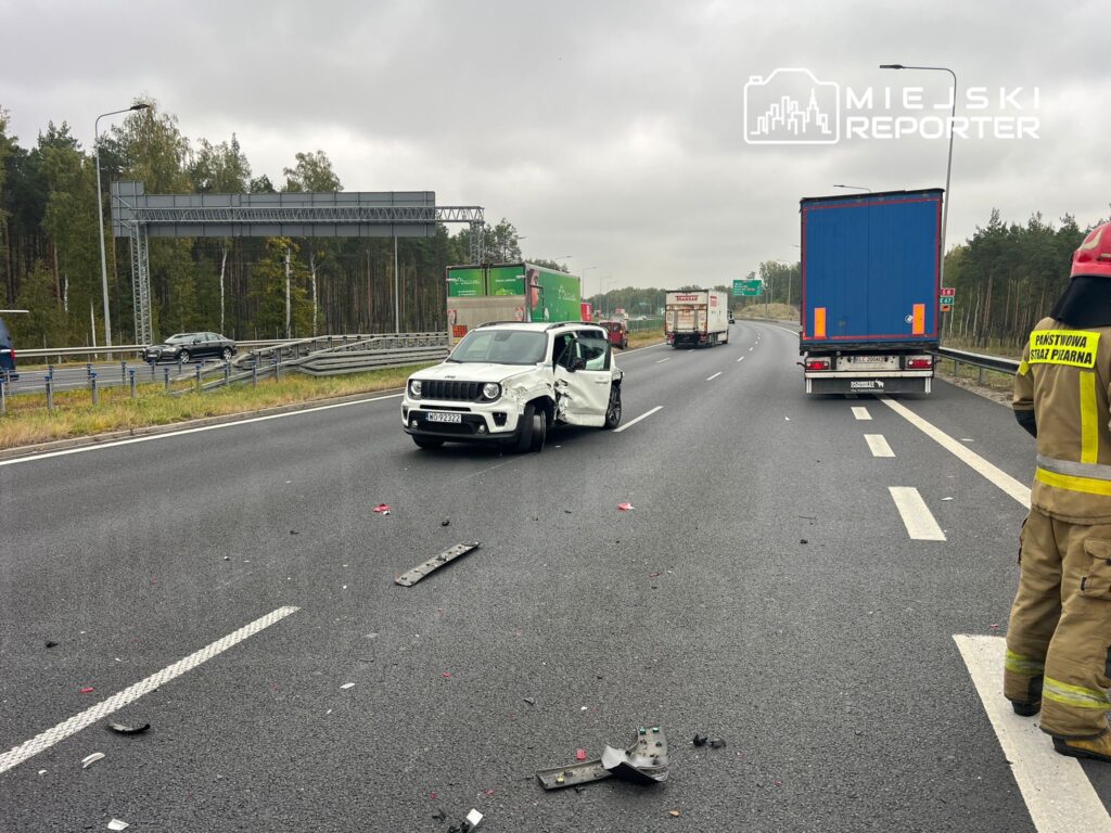 Biały samochód osobowy z otwartymi drzwiami stoi na drodze, obok rozbite pozostałości. W tle widoczne ciężarówki i drzewa.