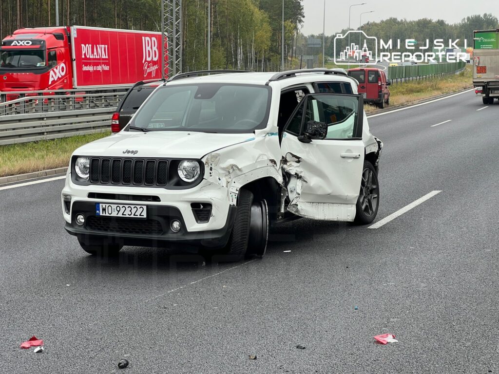 Uszkodzony biały SUV marki Jeep stoi na autostradzie z otwartymi drzwiami, obok widać ciężarówkę w ruchu.