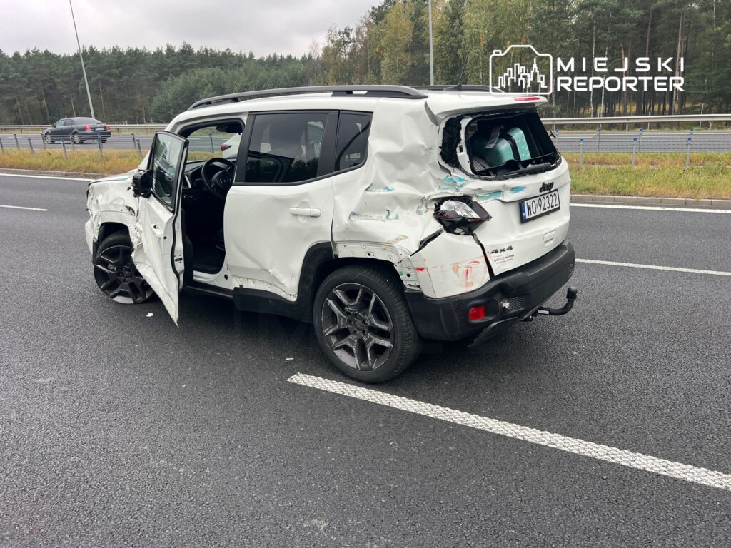 Uszkodzony biały SUV stoi na poboczu drogi, z otwartymi drzwiami i zniszczonym tyłem. W tle widać inne pojazdy.
