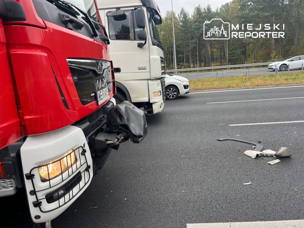 Czerwony ciężarowy pojazd z uszkodzoną przednią częścią stoi obok białego samochodu na drodze, obok leżą pozostałości elementów.