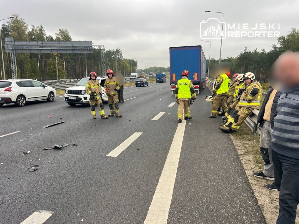 Grupa strażaków w odblaskowych kamizelkach ocenia miejsce wypadku na drodze, obok uszkodzonych pojazdów.