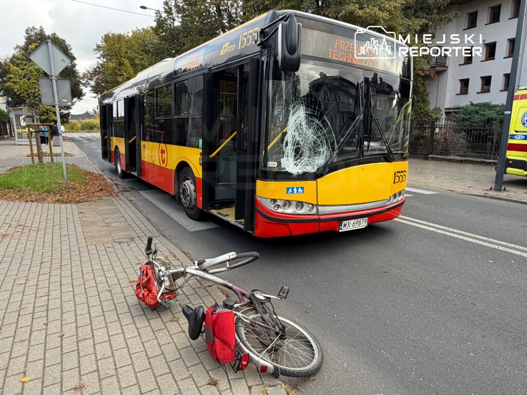 Autobus z uszkodzoną szybą stoi na przystanku, obok leży przewrócony rower z czerwoną torbą.
