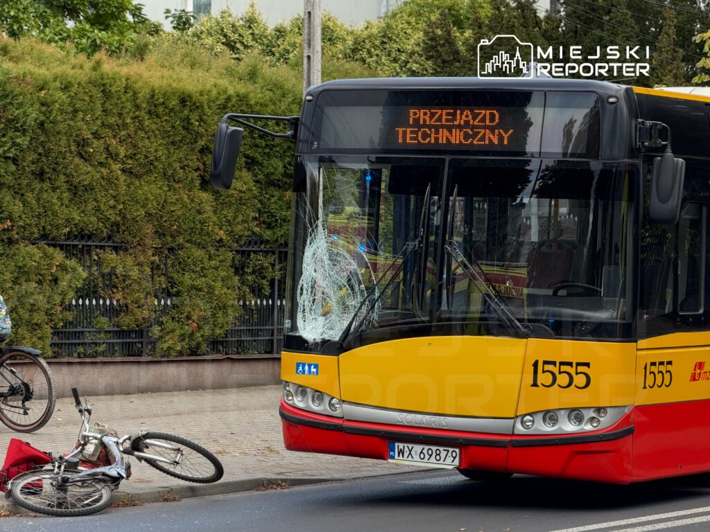 Autobus z uszkodzoną szybą stoi na ulicy obok przewróconego roweru, w tle drzewa i ogrodzenie.