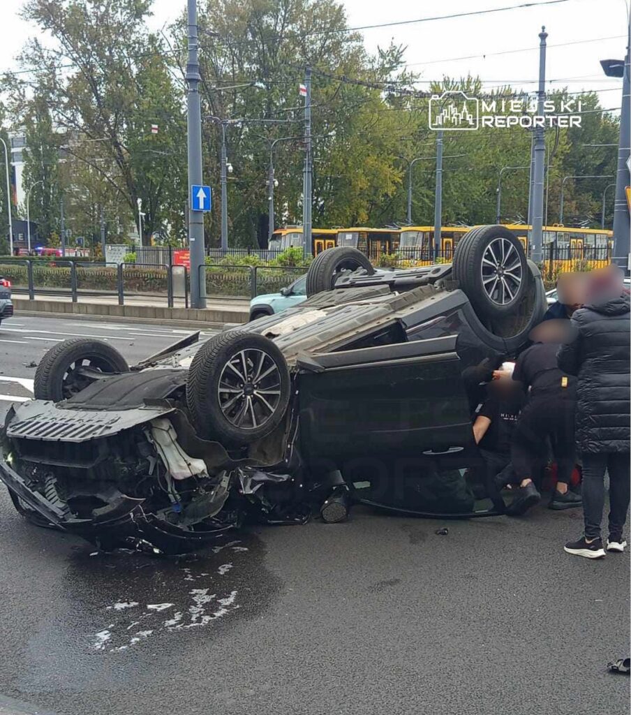 Przewrócony samochód leży na drodze, a kilka osób próbuje udzielić pomocy poszkodowanemu kierowcy.