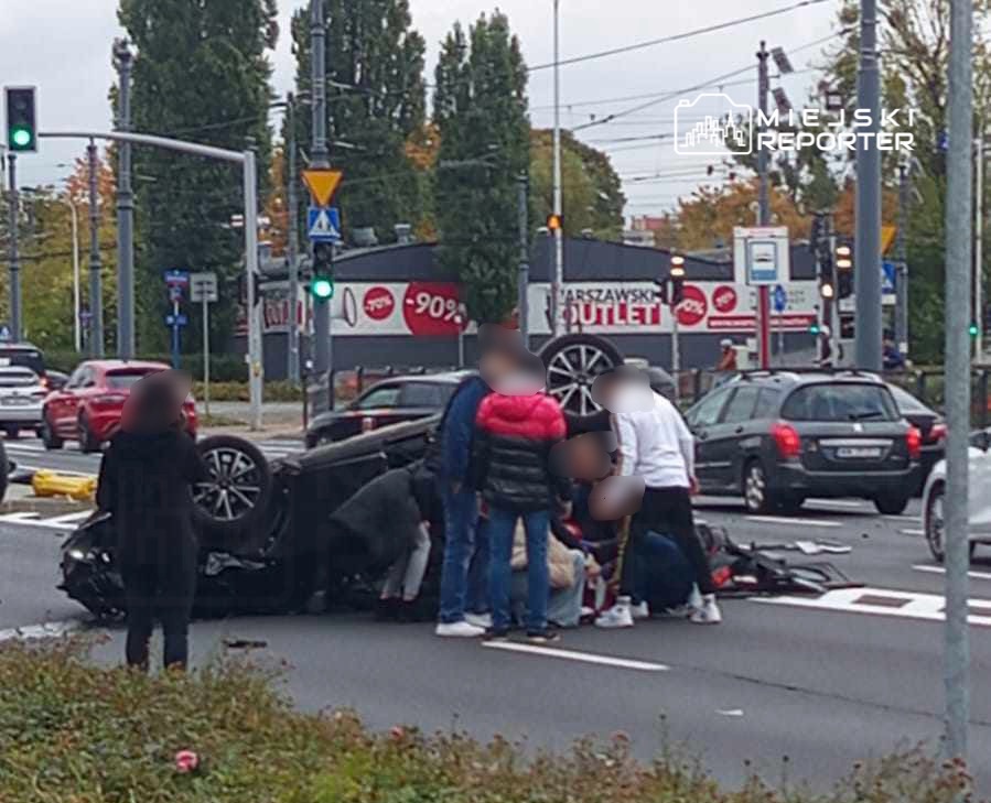 Grupa osób otacza przewrócone auto na skrzyżowaniu, udzielając pomocy poszkodowanemu kierowcy. W tle widać ruch uliczny.