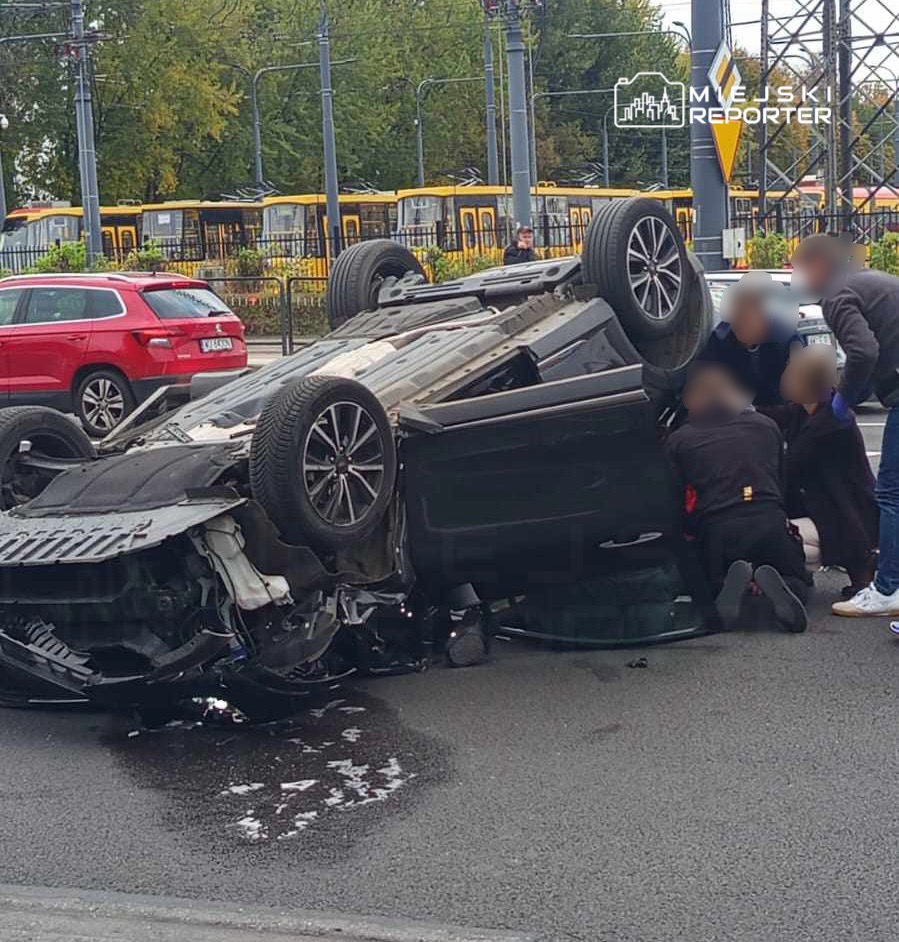 Czarny samochód leży na dachu na jezdni, a kilka osób pomaga poszkodowanemu kierowcy w pobliżu.