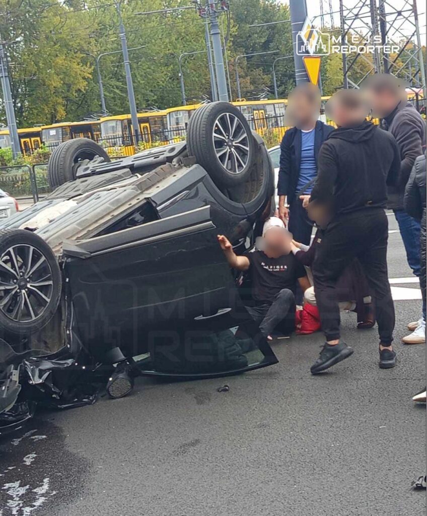 Zdarzenie drogowe z przewróconym samochodem na ulicy, wokół grupka ludzi udzielających pomocy.