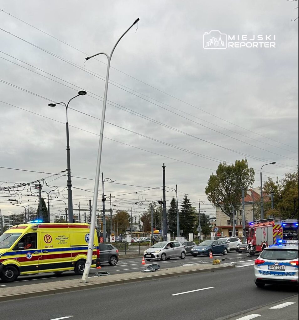 Karetka pogotowia i radiowóz policji zabezpieczają miejsce wypadku na ruchliwej ulicy z uszkodzoną latarnią.