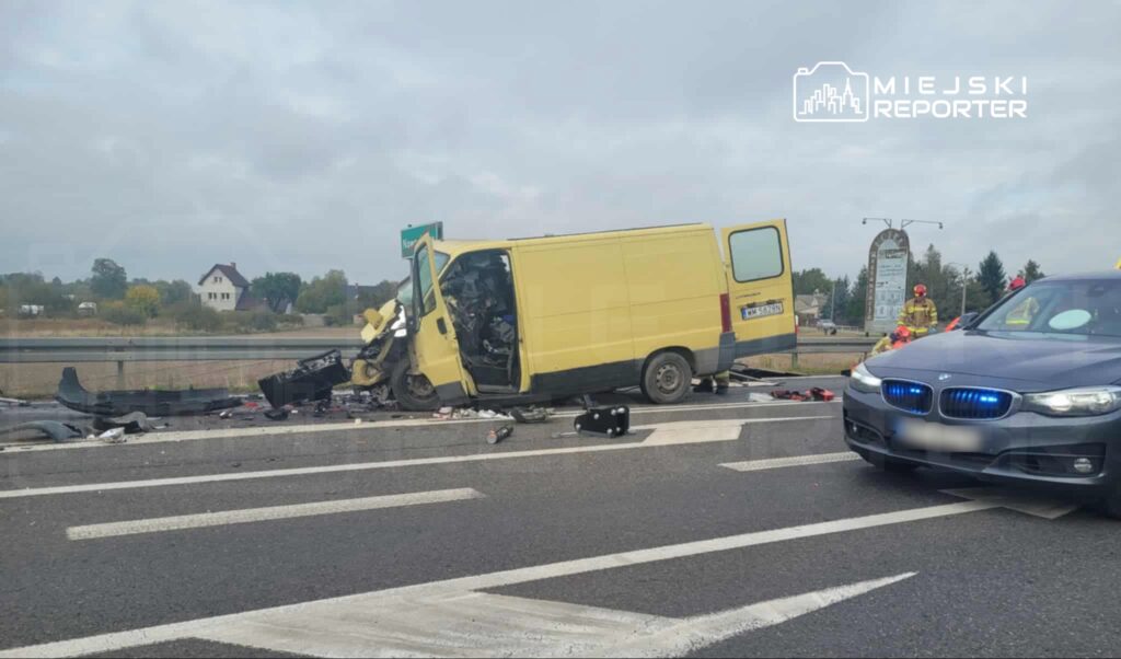 Żółty furgon leży na boku na jezdni, a obok niego widać wrak samochodu oraz interweniujących strażaków.