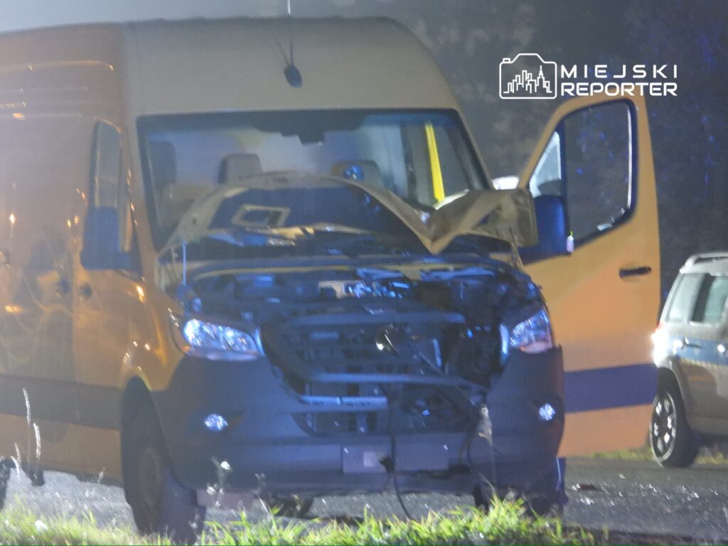 Uszkodzony żółty furgon z otwartą maską stoi na drodze, oświetlony przez niebieskie światła.