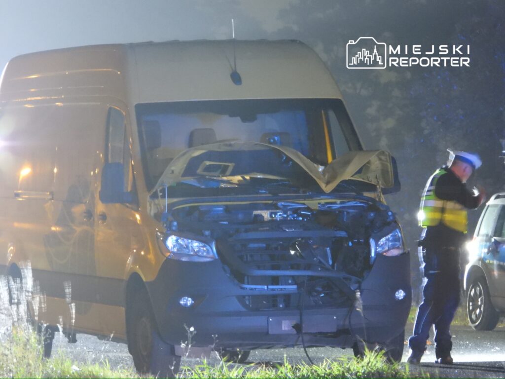 Zniszczona furgonetka z uniesioną maską stoi na drodze, obok policjant w kamizelce odblaskowej przeprowadza czynności.