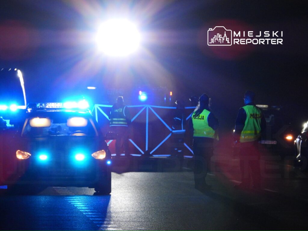 Policjanci w odblaskowych kamizelkach zabezpieczają miejsce zdarzenia w nocy, przy włączonych sygnałach świetlnych.