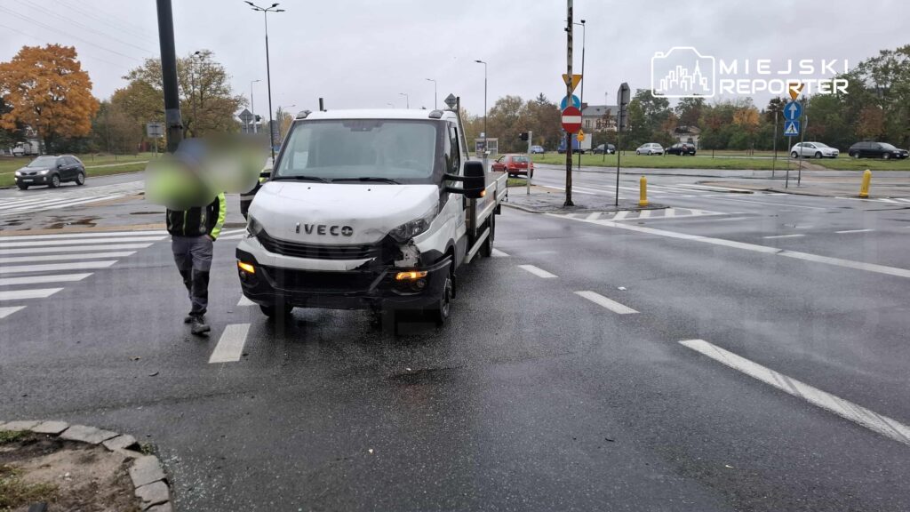 Mężczyzna w odblaskowej kamizelce idzie obok uszkodzonego samochodu dostawczego na mokrej jezdni.