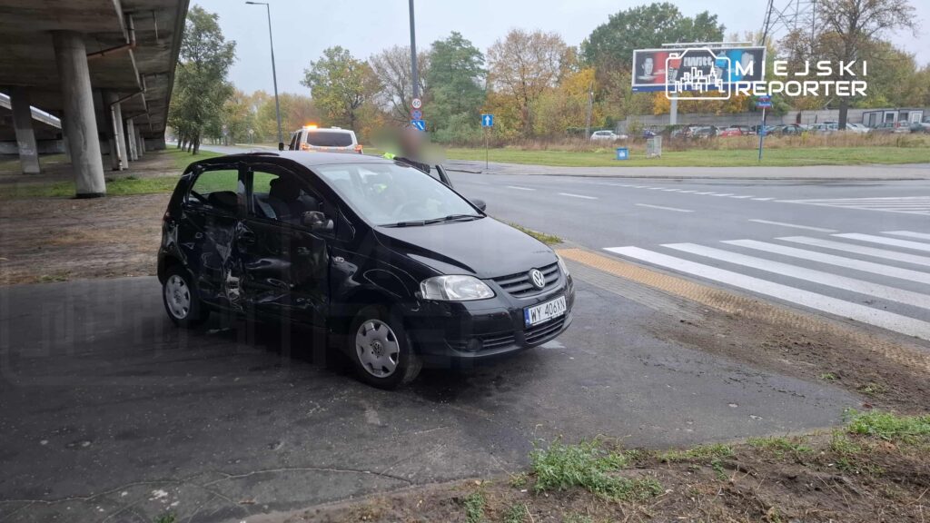 Czarny samochód osobowy z uszkodzeniami stoi na poboczu przy skrzyżowaniu w deszczowej aurze.