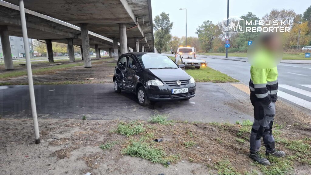 Czarny samochód osobowy z uszkodzeniami stoi pod wiaduktem, obok pracownik w odzieży roboczej obserwuje sytuację.